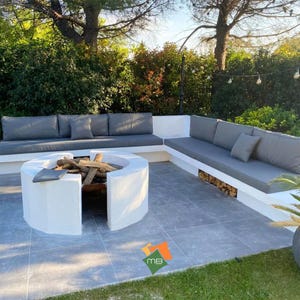 K&ouml;nnte beinhalten: Au&szlig;enbereich mit eingebauter wei&szlig;er Beton-Feuerstelle und grauen Kissen. Die Feuerstelle ist mit Brennholz gef&uuml;llt. Der Sitzbereich hat graue Kissen und Kissen. Die Terrasse ist von Gr&uuml;n umgeben.