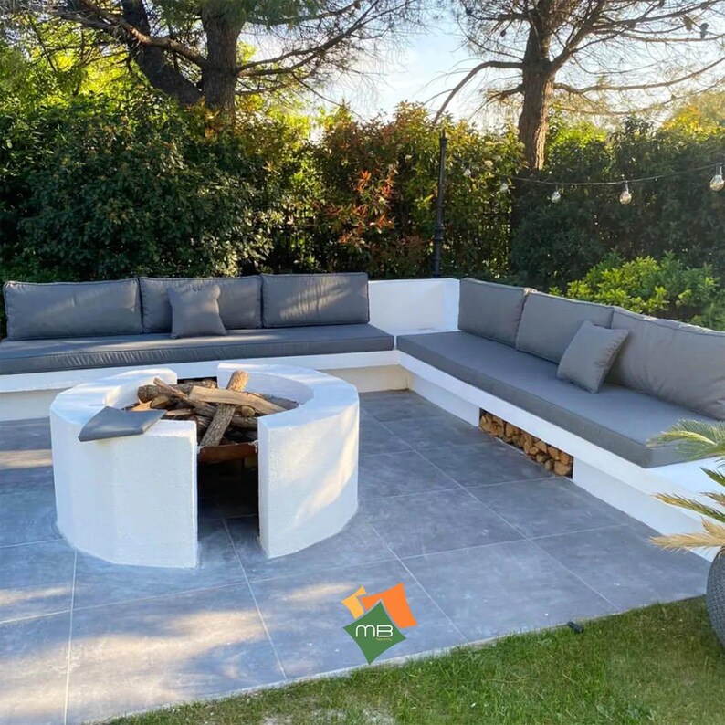 K&ouml;nnte beinhalten: Au&szlig;enbereich mit eingebauter wei&szlig;er Beton-Feuerstelle und grauen Kissen. Die Feuerstelle ist mit Brennholz gef&uuml;llt. Der Sitzbereich hat graue Kissen und Kissen. Die Terrasse ist von Gr&uuml;n umgeben.