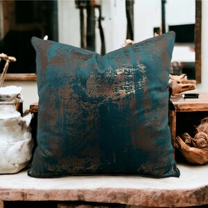 May include: A teal blue throw pillow with a gold metallic abstract pattern. The pillow is sitting on a wooden surface.