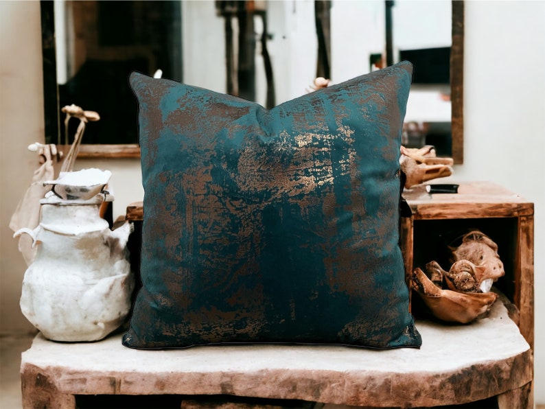 May include: A teal blue throw pillow with a gold metallic abstract pattern. The pillow is sitting on a wooden surface.