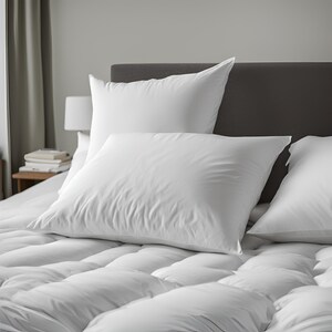 May include: Three white pillows on a white bed with a gray headboard. The pillows are arranged in a stack with the largest pillow on the bottom and the smallest pillow on top.