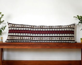 Puede incluir: Una almohada larga y rectangular con fondo blanco y un patrón repetitivo de formas geométricas negras, rojas y doradas. La almohada está sentada en un banco de madera.