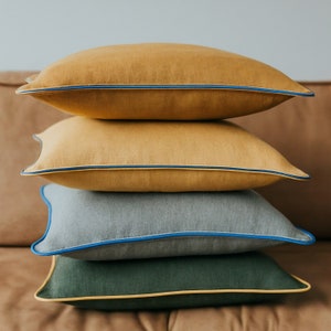 May include: Four decorative pillows stacked on top of each other. The top two pillows are mustard yellow, the third is gray, and the bottom is dark green. Each pillow has a contrasting trim in blue or yellow.