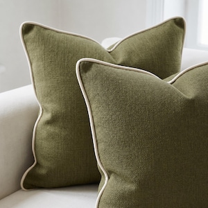 May include: Two square throw cushions in a deep olive green colour with a textured fabric finish. Each cushion features a contrasting cream-coloured piping detail along the edges, providing a clean and defined border to the square shape of the items.