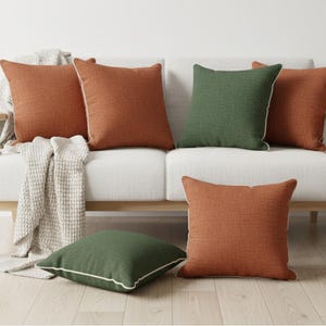 May include: A modern living room scene featuring a white sofa with wooden legs, adorned with several square throw pillows. The pillows are in shades of rust orange and green, with white piping. A neutral-toned throw blanket and a potted plant complete the cozy setting.