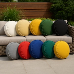 Puede incluir: Una colección de cojines redondos para exteriores en varios colores, incluyendo crema, amarillo, verde y negro, dispuestos sobre un sofá de mimbre. Cojines adicionales en gris, rojo, azul y turquesa se exhiben en el suelo, ofreciendo una vibrante paleta de colores.