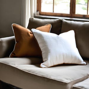 Könnte beinhalten: Zwei quadratische Kissen auf einem Sofa. Eines ist weiß mit braunem Rand, das andere ist braun mit weißem Rand. Die Kissen sind schlicht gestaltet und verleihen dem Wohnzimmer einen Farbtupfer.