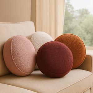 Könnte beinhalten: Vier runde Zierkissen in Rosa-, Creme-, Burgunder- und Orangebrauntönen liegen auf einem beigen Sofa. Die Kissen haben eine strukturierte, flauschige Oberfläche und sind in einer lässigen Gruppe angeordnet, die dem Wohnraum Farbe und Komfort verleiht.