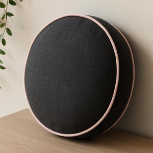 May include: A round, black decorative pillow with a light pink trim. The pillow is made of a textured fabric and is placed on a wooden surface. The pillow is a solid color and has a simple, modern design.