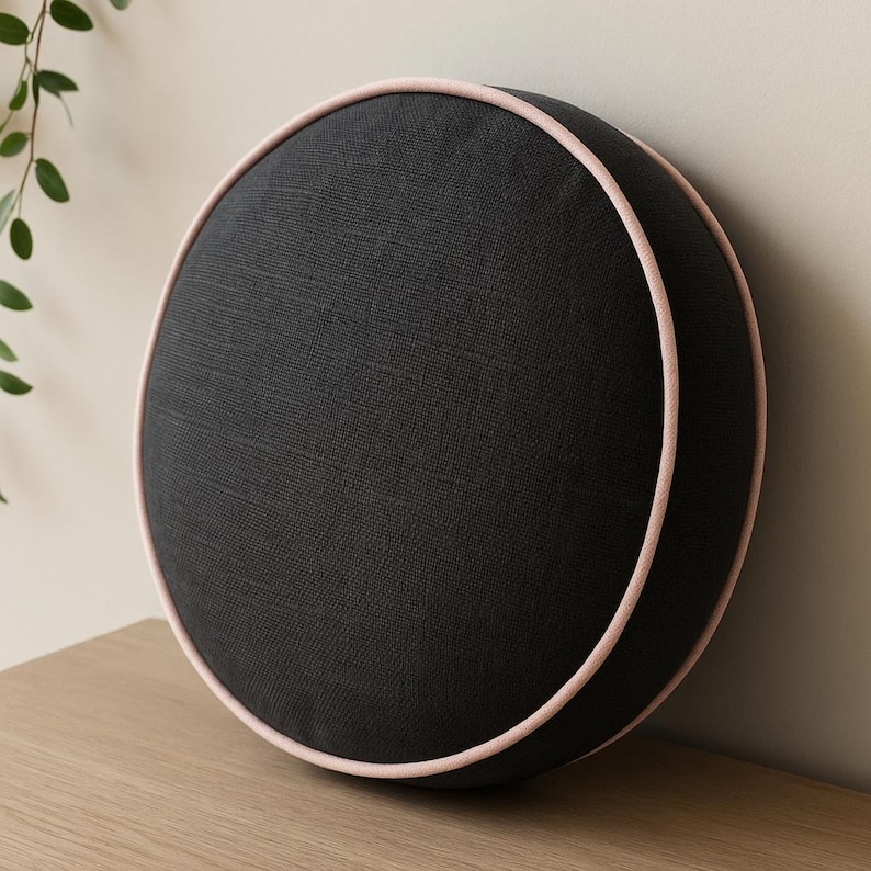 May include: A round, black decorative pillow with a light pink trim. The pillow is made of a textured fabric and is placed on a wooden surface. The pillow is a solid color and has a simple, modern design.