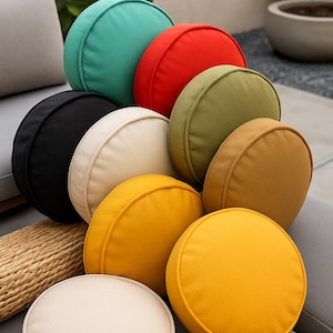 May include: A collection of round, solid-colored throw pillows in various shades including teal, red, olive green, black, cream, tan, and bright yellow. Each cushion features a piped edge and a smooth fabric finish, arranged in a stacked cluster.
