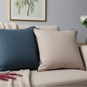 May include: Two square throw pillows on a beige sofa. One pillow is a textured navy blue with black trim, the other is beige with black trim. A beige blanket and a framed picture are in the background.