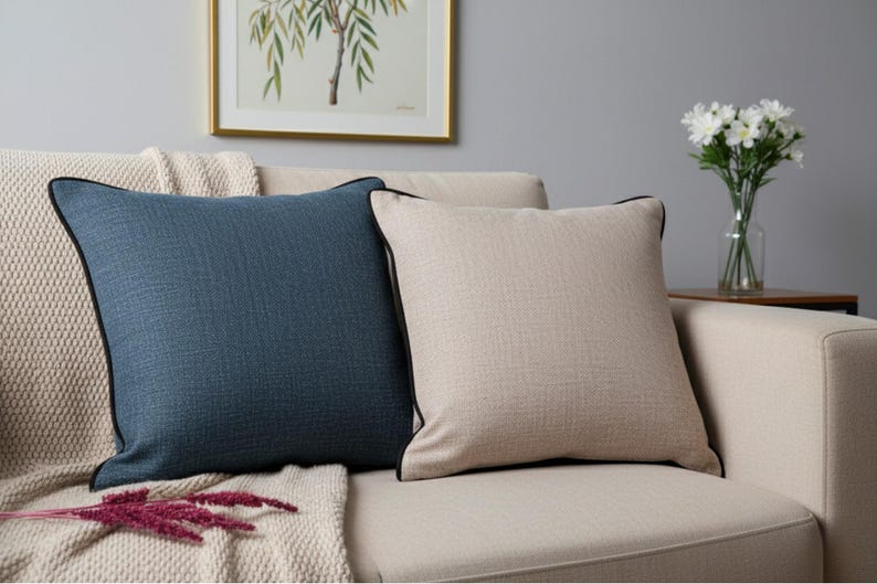 May include: Two square throw pillows on a beige sofa. One pillow is a textured navy blue with black trim, the other is beige with black trim. A beige blanket and a framed picture are in the background.
