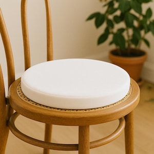 May include: A white, round cushion sits atop a wooden chair with a woven seat. The chair has a curved back and legs, and the wood has a warm, natural finish. The cushion appears to be made of a soft, textured material.
