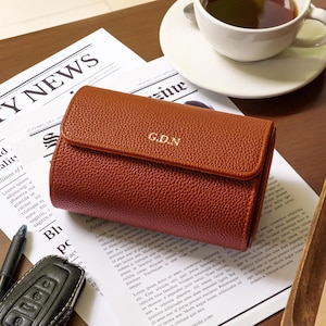 May include: A brown leather wallet with the initials "G.D.N" embossed on the front. The wallet is on a newspaper with the headline "CITY NEWS". A cup of tea and a car key are also visible.