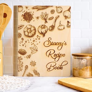 Pode incluir: Um livro de receitas de madeira clara com ilustra&ccedil;&otilde;es gravadas de ervas e especiarias. A capa apresenta o texto "Stacey's Recipe Book" em escrita elegante. Utens&iacute;lios de madeira e uma pequena planta s&atilde;o vis&iacute;veis no fundo.