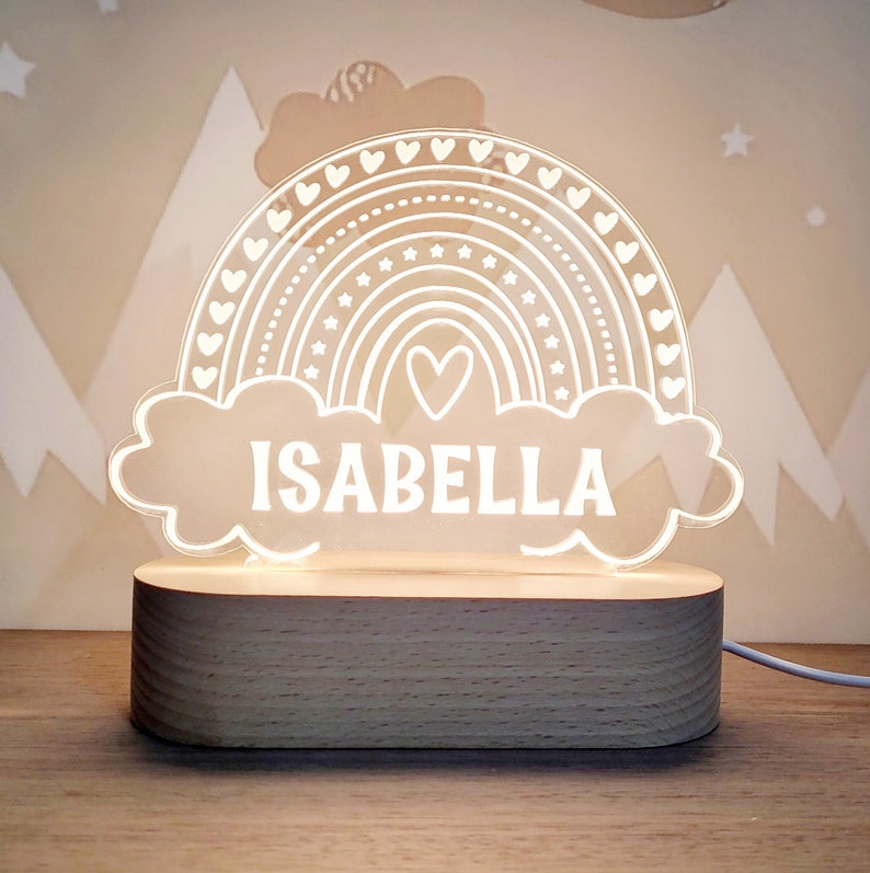 May include: A white acrylic night light shaped like a rainbow with hearts and stars. The rainbow has a small heart in the center and the name "ISABELLA" is written below it. The night light sits on a wooden base.