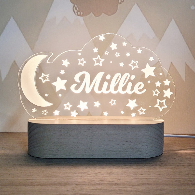 May include: A white acrylic night light shaped like a cloud with stars and a crescent moon. The name "Millie" is written in the center of the cloud. The night light sits on a wooden base.