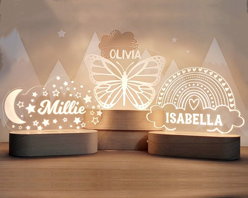 May include: Three wooden night lights with personalized names. The first night light features a cloud with a crescent moon and stars, the second features a butterfly, and the third features a rainbow with a heart. The names Millie, Olivia, and Isabella are written on each night light.