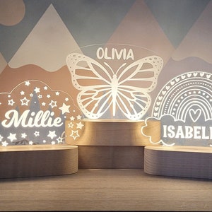 May include: Three wooden night lights with personalized names. The first night light features a white cloud with stars and a crescent moon, the name "Millie" is written inside the cloud. The second night light features a white butterfly with the name "Olivia" written above it. The third night light features a white rainbow with hearts and the name "Isabella" written below it.