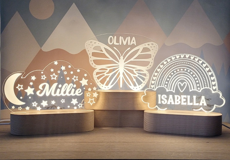 May include: Three wooden night lights with personalized names. The first night light features a white cloud with stars and a crescent moon, the name "Millie" is written inside the cloud. The second night light features a white butterfly with the name "Olivia" written above it. The third night light features a white rainbow with hearts and the name "Isabella" written below it.