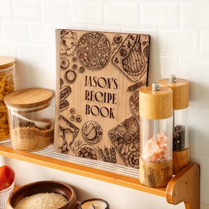 Pode incluir: Um livro de receitas de madeira com o texto "JASON'S RECIPE BOOK" &eacute; exibido em uma prateleira de madeira. A capa apresenta ilustra&ccedil;&otilde;es gravadas de alimentos como pizza e bife. Outros itens de cozinha incluem potes de vidro.
