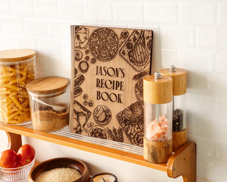 Pode incluir: Um livro de receitas de madeira com o texto "JASON'S RECIPE BOOK" &eacute; exibido em uma prateleira de madeira. A capa apresenta ilustra&ccedil;&otilde;es gravadas de alimentos como pizza e bife. Outros itens de cozinha incluem potes de vidro.