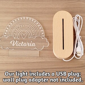 May include: A white acrylic rainbow night light with the name "Victoria" inscribed in the cloud. The light is plugged into a wooden base with a USB cord.