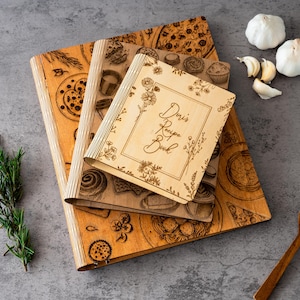 Pode incluir: Livros de receitas de madeira com ilustra&ccedil;&otilde;es de comida gravadas e o texto "Doris Recipe Book". Os livros s&atilde;o castanhos claros com um design de borda floral. Alecrim fresco, alho e uma colher de pau tamb&eacute;m est&atilde;o presentes.