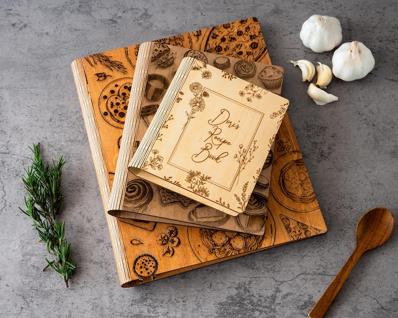 Pode incluir: Livros de receitas de madeira com ilustra&ccedil;&otilde;es de comida gravadas e o texto "Doris Recipe Book". Os livros s&atilde;o castanhos claros com um design de borda floral. Alecrim fresco, alho e uma colher de pau tamb&eacute;m est&atilde;o presentes.