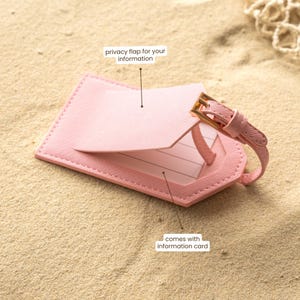 Puede incluir: Una etiqueta de equipaje rosa con una solapa de privacidad y una correa con una hebilla dorada. La etiqueta est&aacute; sobre una superficie arenosa. El texto en la etiqueta dice "privacy flap for your information" y "comes with information card."
