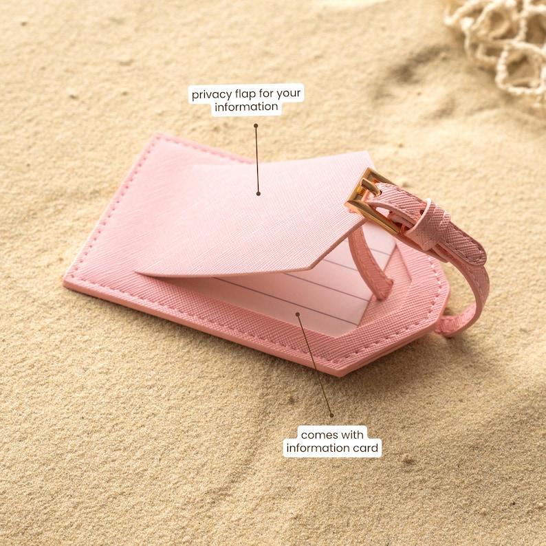 Puede incluir: Una etiqueta de equipaje rosa con una solapa de privacidad y una correa con una hebilla dorada. La etiqueta est&aacute; sobre una superficie arenosa. El texto en la etiqueta dice "privacy flap for your information" y "comes with information card."