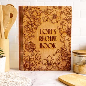Pode incluir: Um livro de receitas de madeira com gravuras florais e o texto "LORI'S RECIPE BOOK". O livro &eacute; rodeado por utens&iacute;lios de madeira, uma pequena planta em vaso e frascos de vidro cheios de ingredientes.