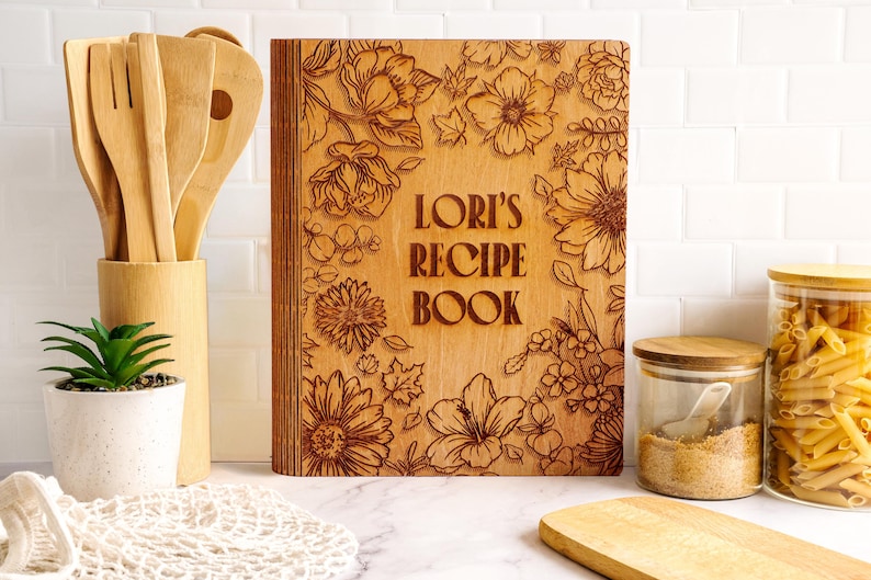 Pode incluir: Um livro de receitas de madeira com gravuras florais e o texto "LORI'S RECIPE BOOK". O livro &eacute; rodeado por utens&iacute;lios de madeira, uma pequena planta em vaso e frascos de vidro cheios de ingredientes.