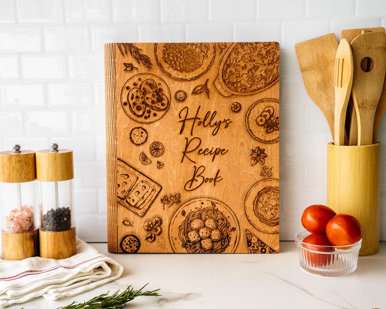 Pode incluir: Um livro de receitas de madeira com ilustra&ccedil;&otilde;es gravadas de alimentos, incluindo massas e pizza. A capa diz "Holly's Recipe Book". Moedores de sal e pimenta, utens&iacute;lios de madeira e tomates tamb&eacute;m s&atilde;o vis&iacute;veis.