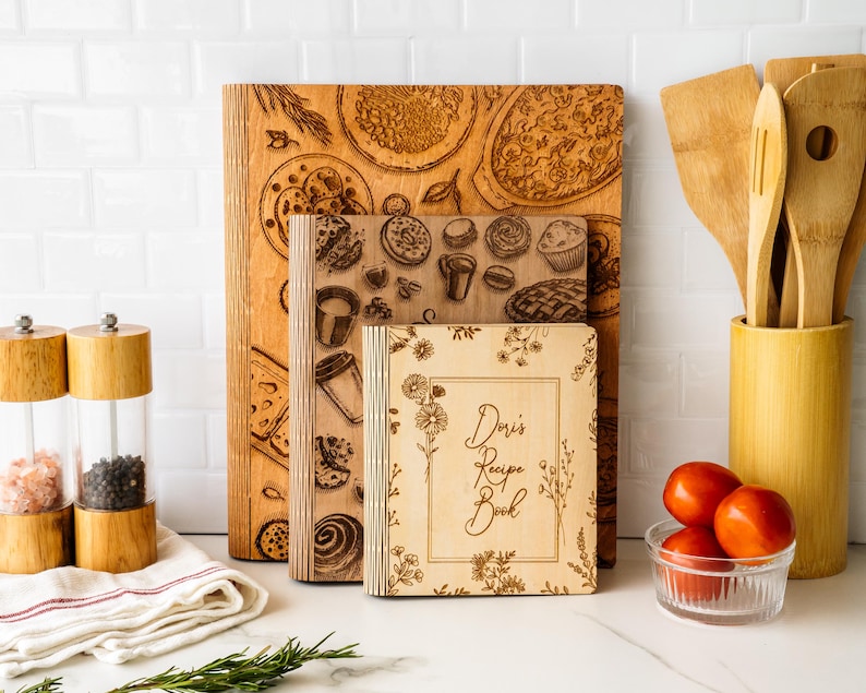 Pode incluir: Tr&ecirc;s livros de receitas e t&aacute;buas de corte de madeira com desenhos de comida gravados. O livro menor &eacute; rotulado "Doris Recipe Book" com detalhes florais. Utens&iacute;lios de cozinha, tomates e saleiros e pimenteiros tamb&eacute;m s&atilde;o vis&iacute;veis.