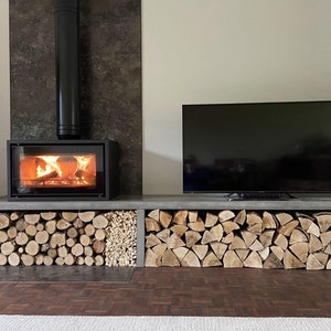 May include: A modern fireplace with a black metal chimney and a glass door, set into a wall with a dark gray tile. The fireplace is surrounded by a concrete shelf with stacked firewood.
