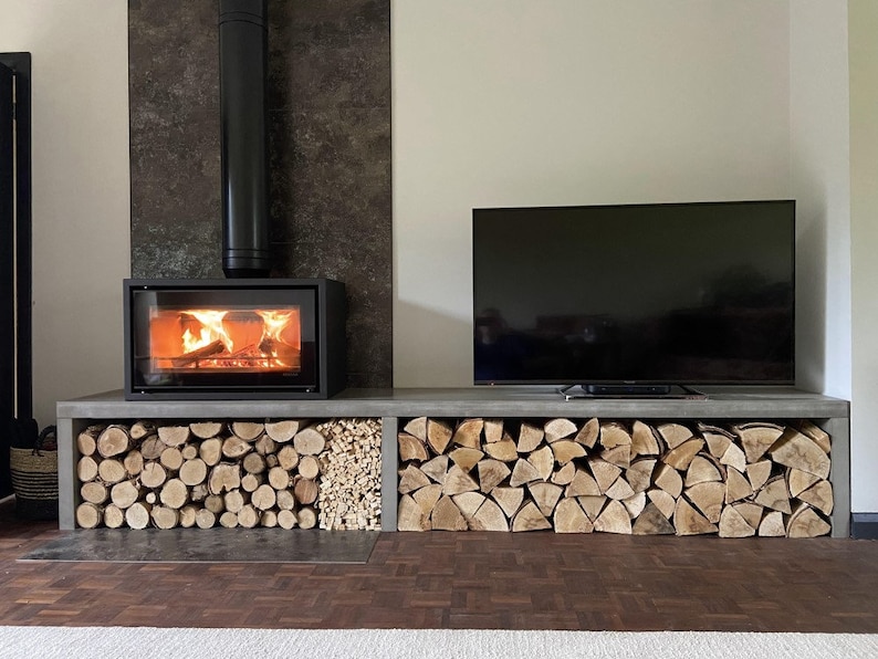 May include: A modern fireplace with a black metal chimney and a glass door, set into a wall with a dark gray tile. The fireplace is surrounded by a concrete shelf with stacked firewood.