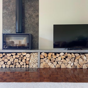 May include: A modern fireplace with a black metal chimney and a black glass door. The fireplace is built into a wall with a gray tile surround. A flat screen TV is mounted above a concrete shelf with stacked firewood.