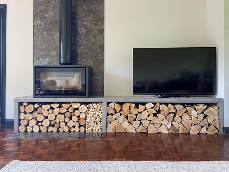 May include: A modern fireplace with a black metal chimney and a black glass door. The fireplace is built into a wall with a gray tile surround. A flat screen TV is mounted above a concrete shelf with stacked firewood.