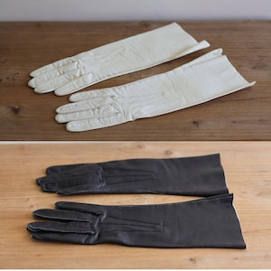 Könnte beinhalten: Zwei Paar lange Handschuhe, einer in Elfenbein und einer in Schwarz, auf einer Holzoberfläche. Die Handschuhe scheinen aus Leder zu sein, mit einem klassischen, eleganten Design. Die Handschuhe sind lang und reichen den Arm hinauf.