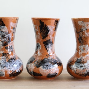 Könnte beinhalten: Drei Keramikvasen mit bauchigem Boden und ausgestelltem Rand. Die Vasen sind in einem gesprenkelten Muster aus Orange, Schwarz, Weiß und Grau glasiert. Die Vasen werden auf einer Holzoberfläche ausgestellt.