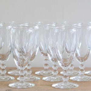 Pode incluir: Conjunto de taças de vinho transparentes. Cada taça apresenta um design texturizado e uma haste com contas. As taças estão dispostas sobre uma superfície de madeira, para uma mesa elegante.