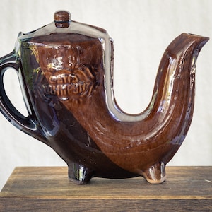 Könnte beinhalten: Eine braune Teekanne aus Keramik mit einem einzigartigen Ausguss in Form eines gebogenen Horns. Die Teekanne hat einen Deckel und einen Griff. Die Teekanne steht auf einem Holzsockel. Die Teekanne hat die Worte "CAT & CHIMP" auf der Seite eingeprägt.