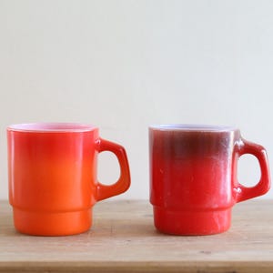 Puede incluir: Dos tazas de vidrio vintage con un esquema de color degradado. Una taza hace la transición de naranja a rojo, la otra de rojo a un tono más oscuro. Ambas tienen un asa y una base apilada, sobre una superficie de madera.