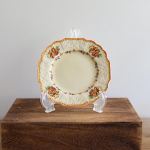 Peut inclure: Une petite assiette en céramique de couleur crème avec un motif floral en relief dans des tons d'orange, jaune et bleu. L'assiette est sur un support en bois.