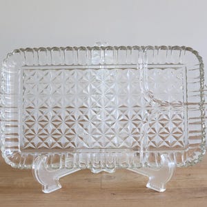 Może przedstawiać: Przezroczysta szklana taca do serwowania z diamentowym wzorem i trzema podzielonymi sekcjami. Taca ma ozdobny falisty brzeg i jest podtrzymywana przez trzy przezroczyste plastikowe nóżki.