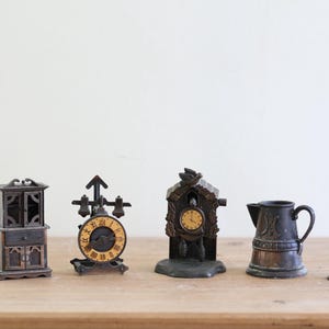 May include: A collection of miniature antique items, including a cabinet, two clocks, a pitcher, and a small weight. The clocks have yellow clock faces. The items are made of metal and sit on a wooden surface.