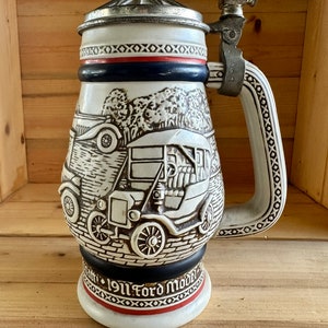 May include: A vintage ceramic beer stein with a detailed relief of a 1911 Ford Model T. The stein features a hinged metal lid, a large handle, and a decorative band with text. The color scheme includes white, black, red, and silver accents.