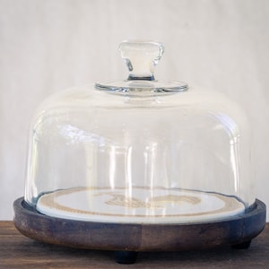 Peut inclure: Une cloche en verre transparent avec une base en bois et une assiette blanche avec un motif doré à l'intérieur. La cloche est sur une table en bois.